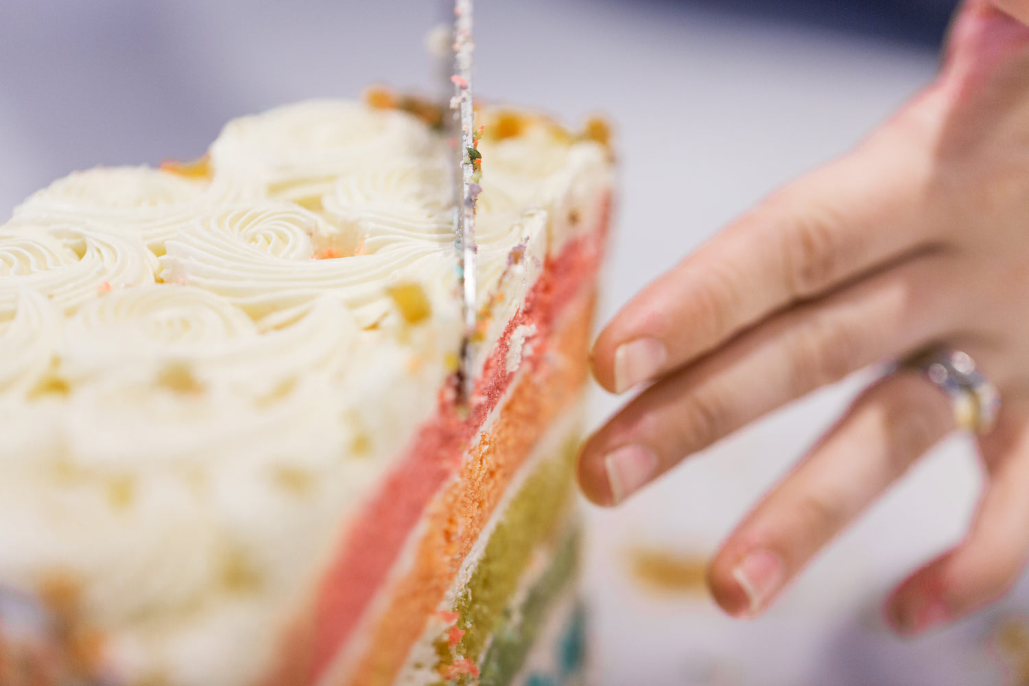 Eine Nahaufnahme eines angeschnittenen Stücks einer Regenbogentorte mit mehreren bunten Schichten, bedeckt mit weißer Buttercreme. Im Vordergrund ist eine Hand mit einem Ring zu sehen, die das Stück beim Schneiden hält.