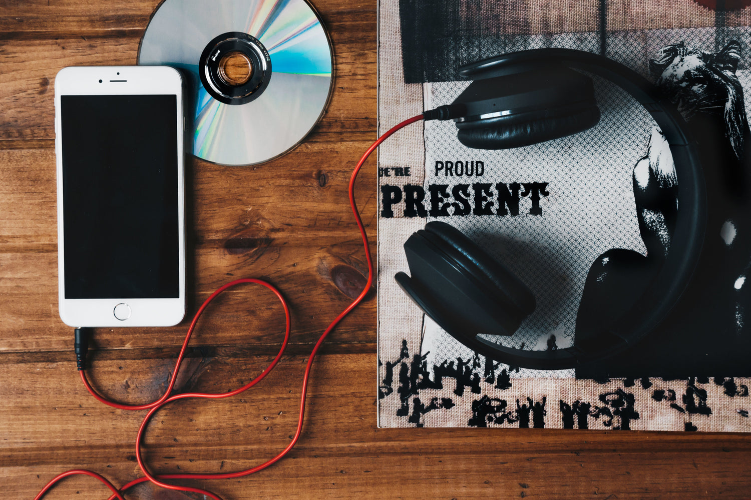 Links ein Smartphone mit schwarzem Bildschirm und angeschlossenem roten Kopfhörerkabel, daneben eine schimmernde CD. Rechts im Bild ein schwarzer Over-Ear-Kopfhörer, der auf einem bedruckten Stoff mit der Aufschrift 'BE PROUD PRESENT' liegt.