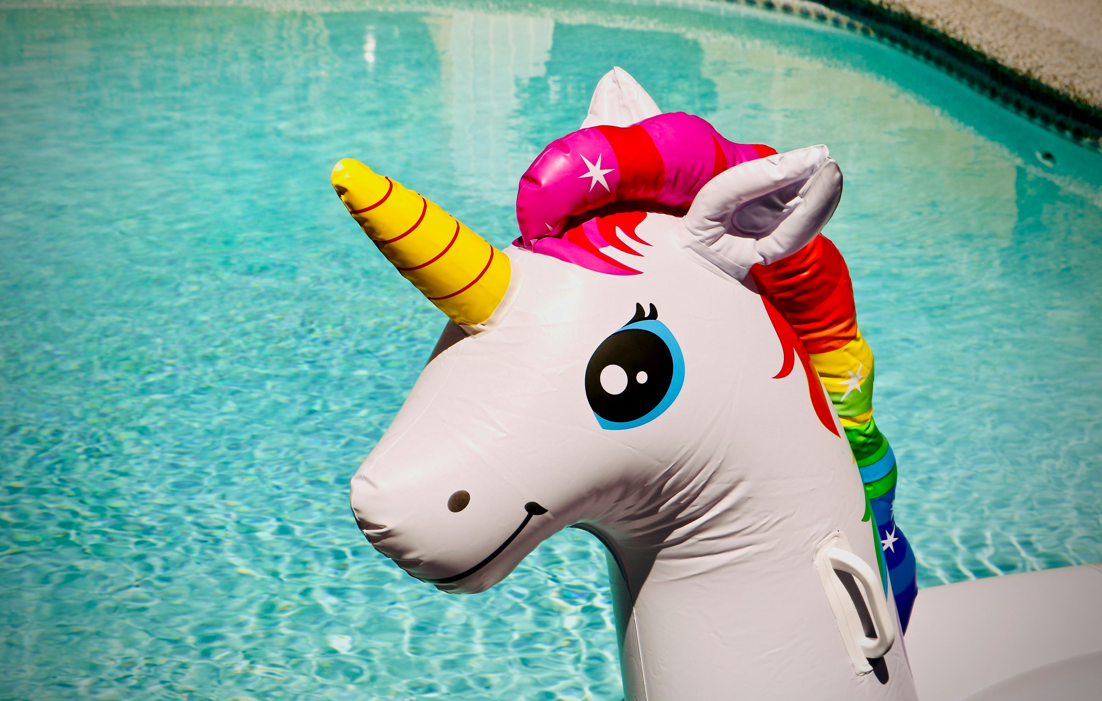 Aufblasbares Einhorn-Schwimmtier in einem Pool mit klarem, türkisfarbenem Wasser. Das Einhorn hat bunte, regenbogenfarbene Mähne, ein gelbes Horn mit roten Streifen und ein fröhliches Gesicht mit großen, leuchtenden Augen.