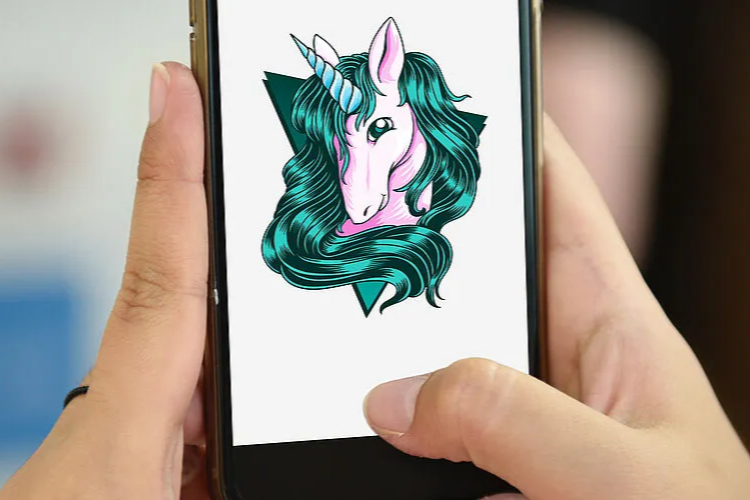 Zwei Hände halten ein Smartphone, auf dessen Bildschirm ein farbenfrohes Einhorn-Illustration zu sehen ist. Das Einhorn hat eine rosa Schnauze, eine grüne Mähne und ein Horn, vor einem weißen Hintergrund.