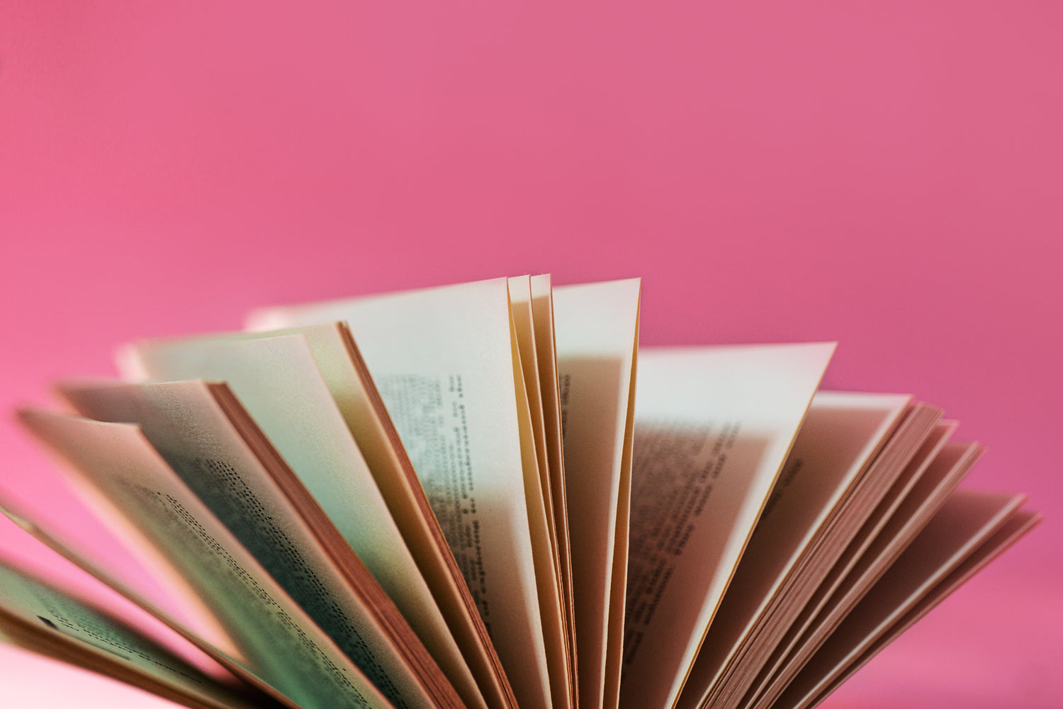 Ein aufgeschlagenes Buch mit leicht bewegten Seiten vor einem kräftigen pinkfarbenen Hintergrund. Die Seiten sind teilweise verschwommen, was eine dynamische und moderne Ästhetik vermittelt.