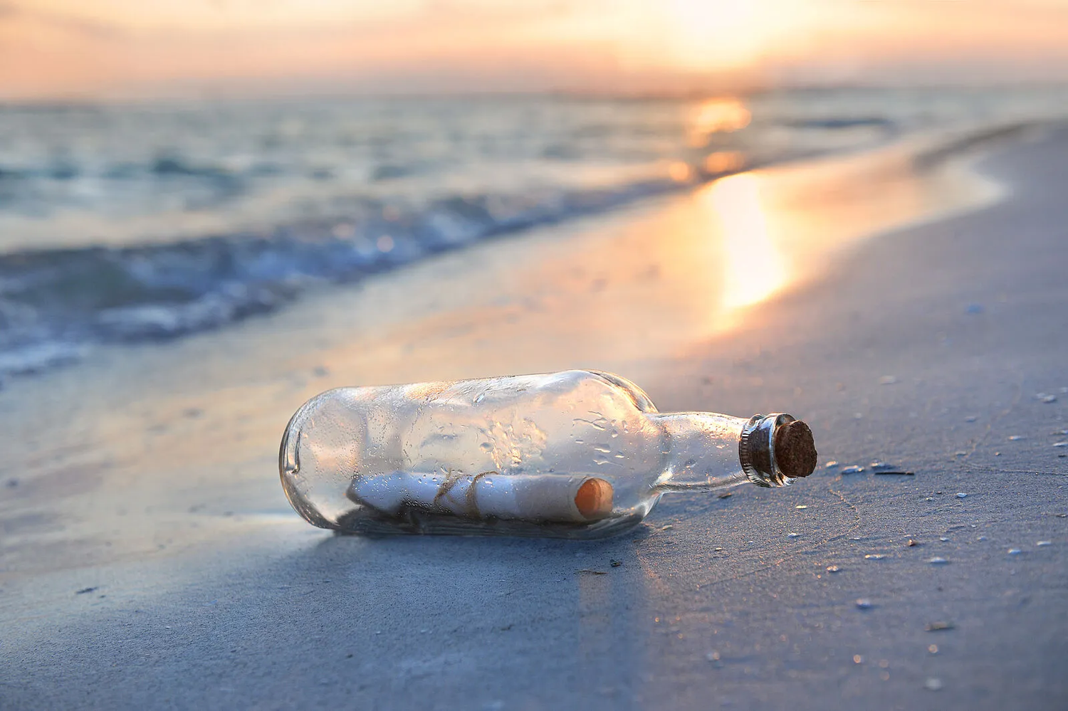 Eine Flaschenpost die am Strand liegt. Im Hintergrund sieht man das Meer und einen Sonnenuntergang.