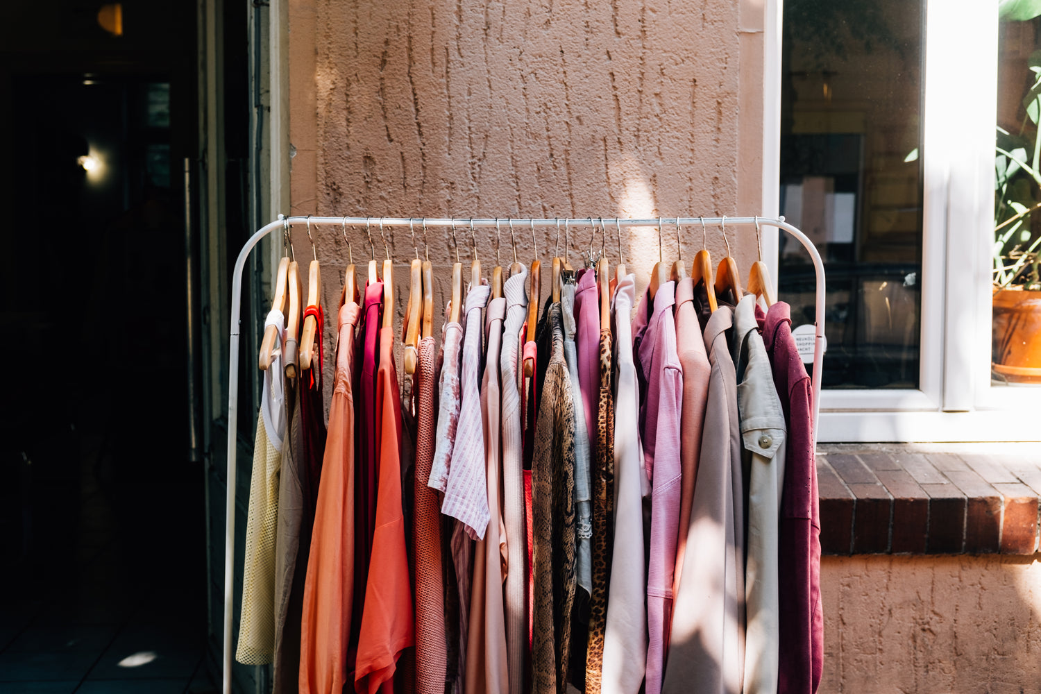 Ein Kleiderständer mit verschiedenen Kleidungsstücken in warmen Farbtönen wie Rosa, Rot, Beige und Braun, der vor einer beigen Hauswand steht. Sonnenlicht fällt auf einige der Kleidungsstücke, während der Hintergrund teils im Schatten liegt.