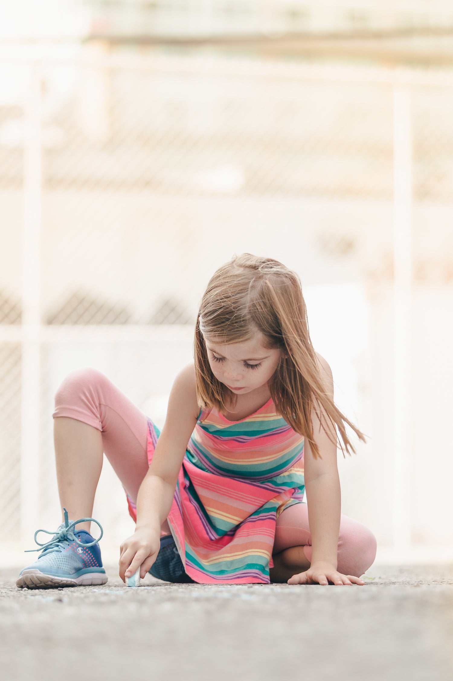 Ein Kind kniet auf dem Boden eines sonnigen Spielplatzes und malt mit einem Stück Kreide auf den Asphalt. Es trägt ein bunt gestreiftes Kleid, pinke Leggings und blaue Turnschuhe. Im Hintergrund ist ein Maschendrahtzaun leicht unscharf zu erkennen.