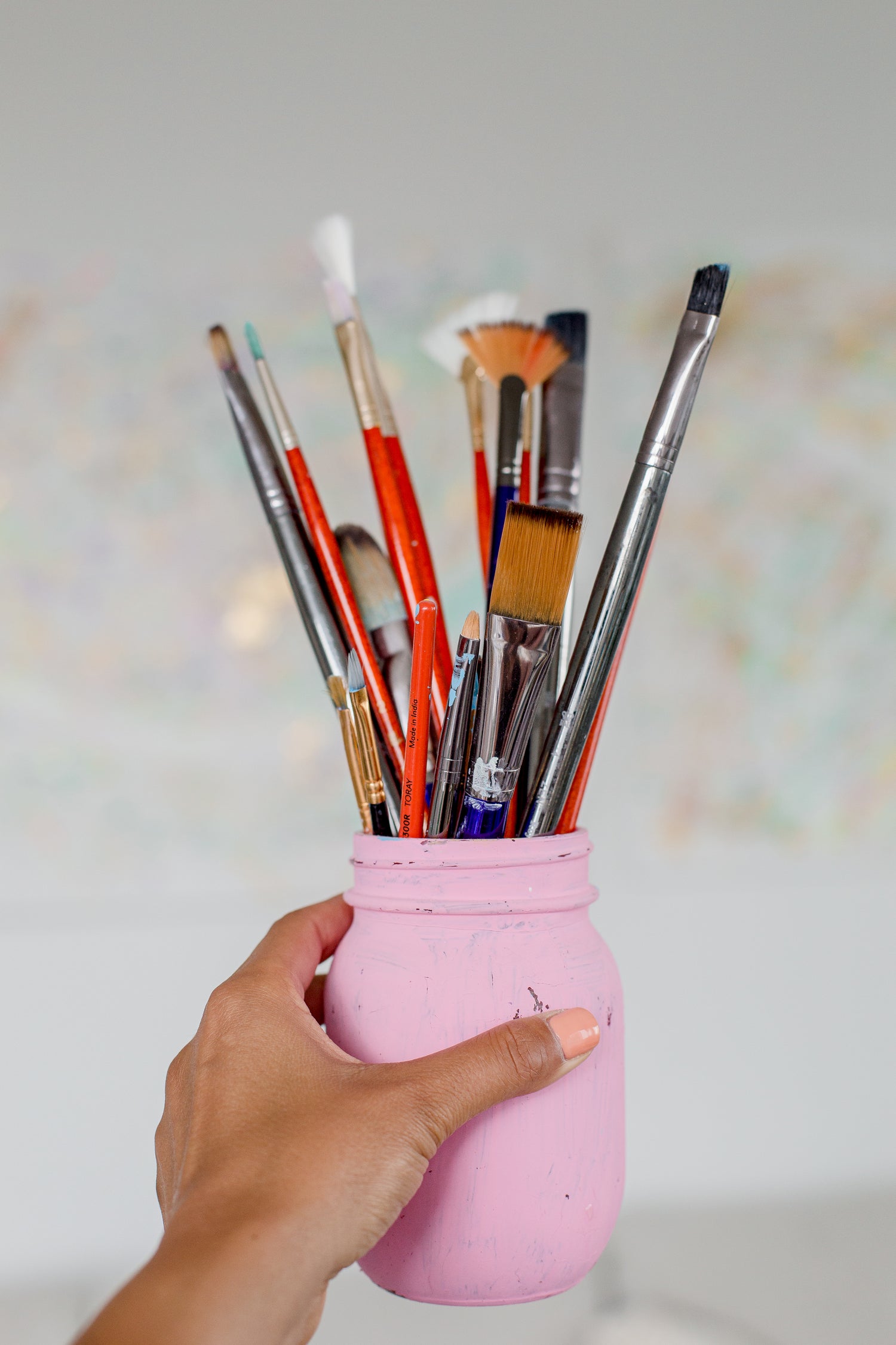 Eine Hand hält ein rosa Einmachglas, das mit verschiedenen Malpinseln gefüllt ist. Die Pinsel haben unterschiedliche Größen und Formen, darunter flache und runde Borsten. Der Hintergrund ist unscharf und zeigt pastellfarbene Muster