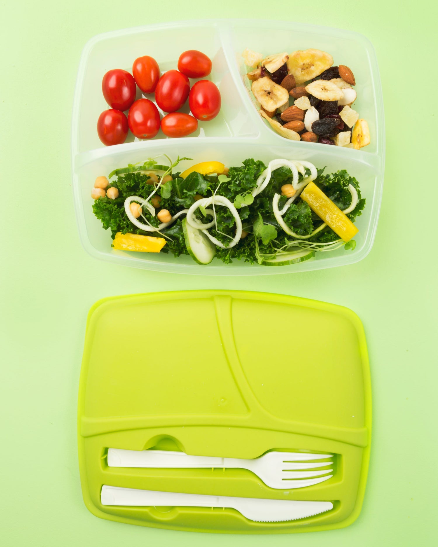 Ein grüner, wiederverwendbarer Bento-Box-Behälter mit drei Fächern, gefüllt mit frischen Lebensmitteln: Kirschtomaten, einer Mischung aus getrockneten Früchten und Nüssen sowie einem grünen Salat mit Kichererbsen, Gurkenscheiben, Zwiebeln und gelben Paprikastreifen. Darunter liegt der passende grüne Deckel mit integriertem weißen Kunststoffbesteck (Messer und Gabel). Der Hintergrund ist hellgrün.