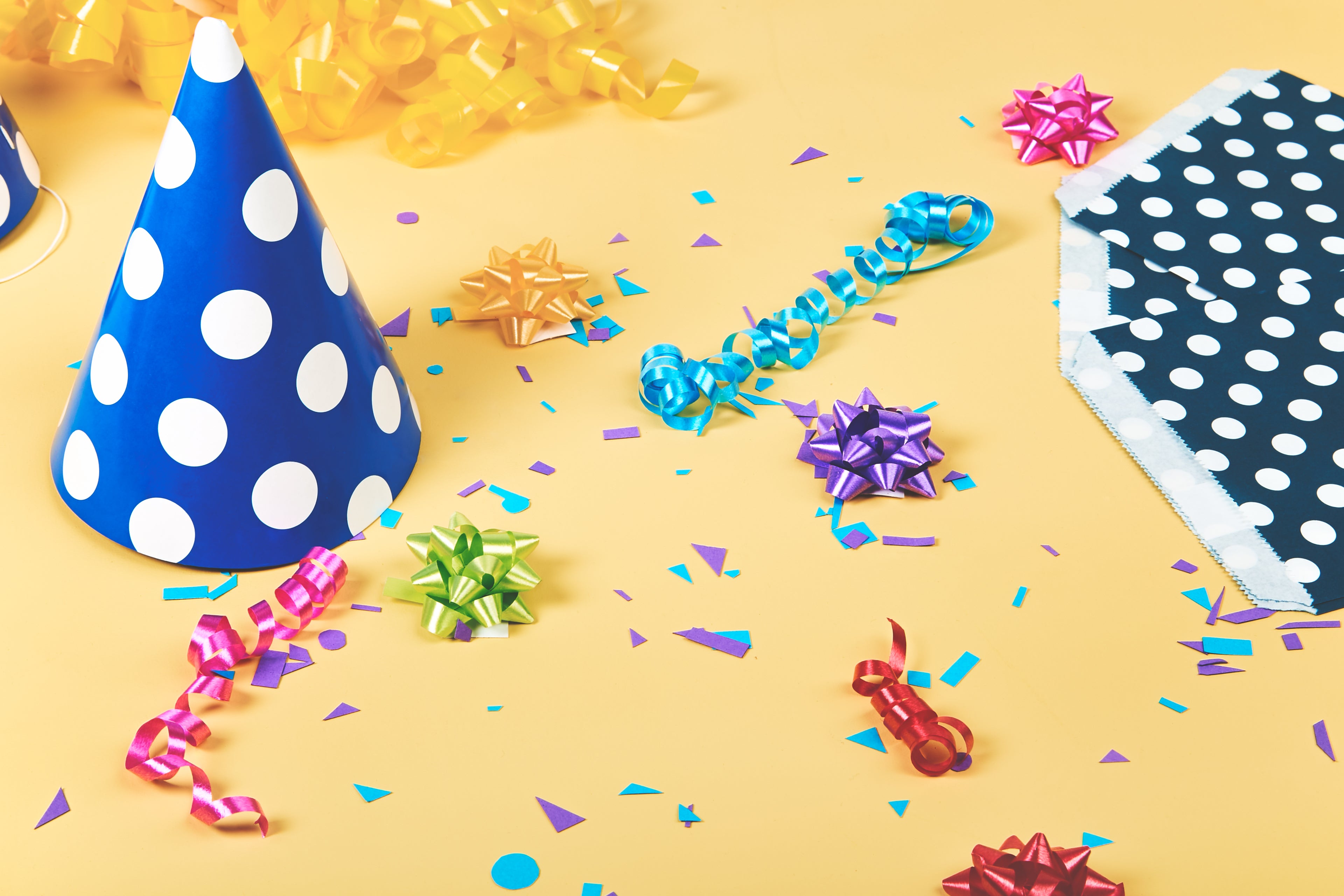 Bunte Partydekoration mit Partyhut, Schleifen, Konfetti und Geschenkbändern auf gelbem Hintergrund.