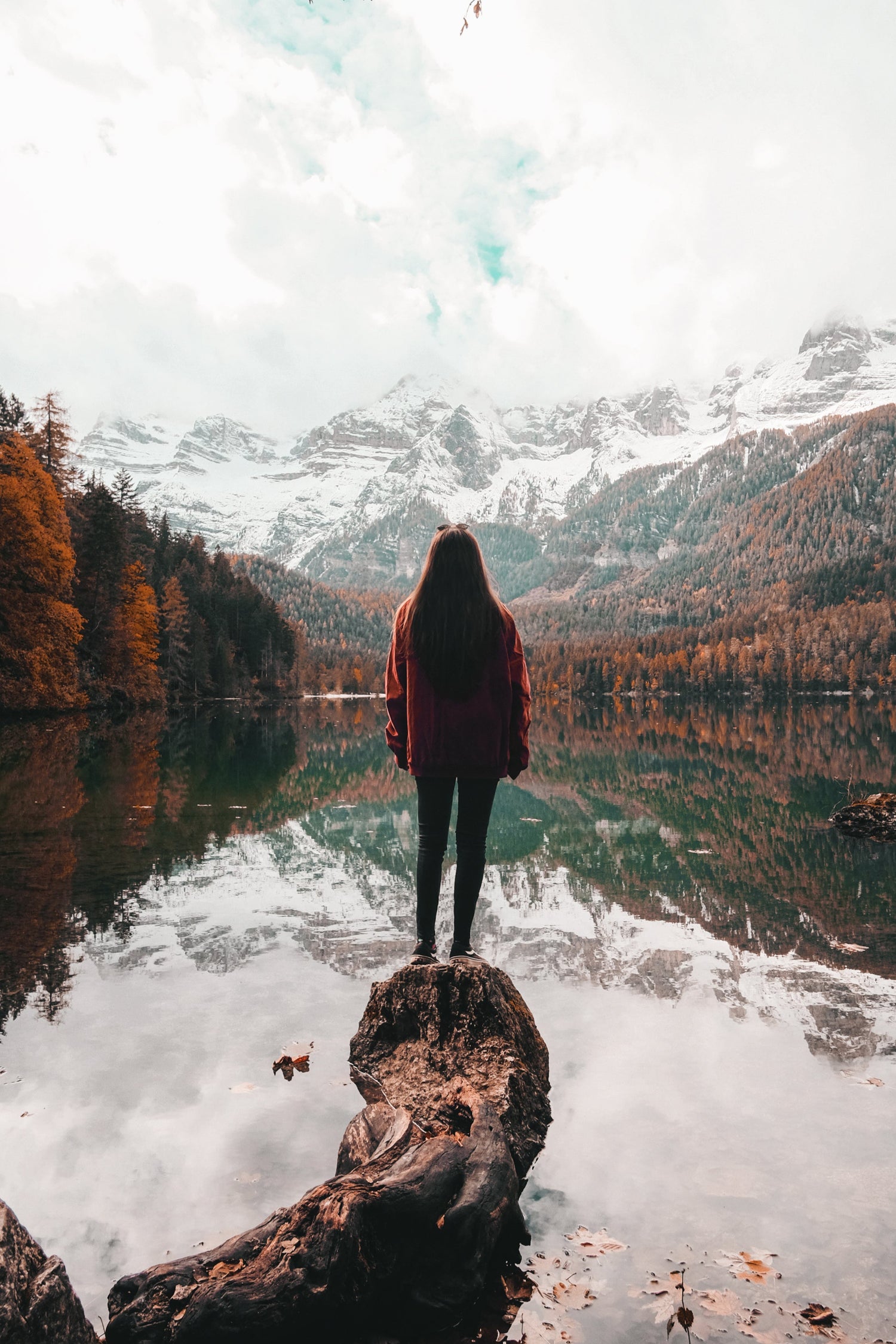 Eine Frau steht auf einem Baumstamm, der in einen ruhigen Bergsee ragt. Sie trägt eine rote Jacke und blickt auf die herbstliche Landschaft mit bunten Bäumen und schneebedeckten Bergen im Hintergrund, die sich im klaren Wasser spiegeln. Der Himmel ist bewölkt, und einige Blätter hängen von einem Ast im oberen Bildrand.