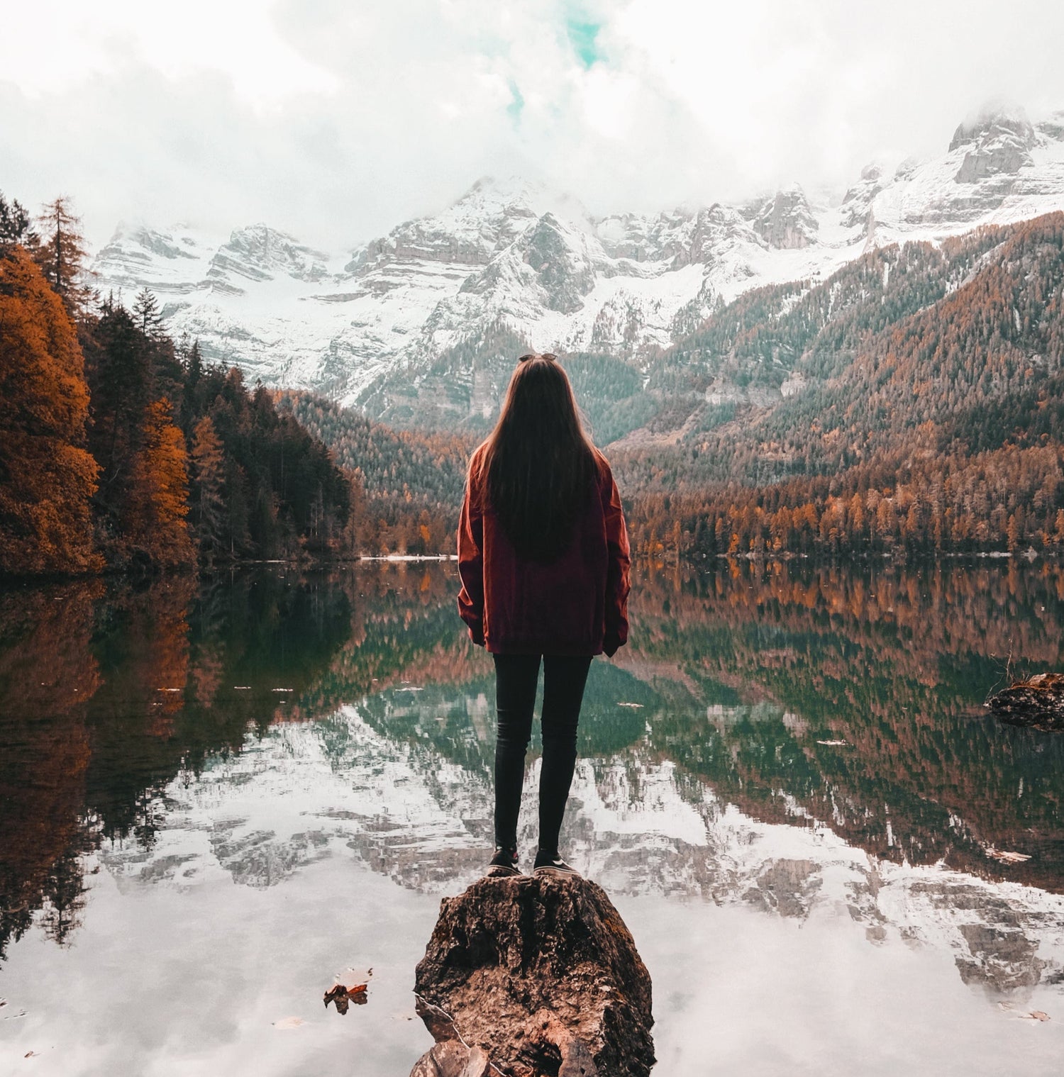 Eine Frau steht auf einem Baumstamm, der in einen ruhigen Bergsee ragt. Sie trägt eine rote Jacke und blickt auf die herbstliche Landschaft mit bunten Bäumen und schneebedeckten Bergen im Hintergrund, die sich im klaren Wasser spiegeln. Der Himmel ist bewölkt, und einige Blätter hängen von einem Ast im oberen Bildrand.