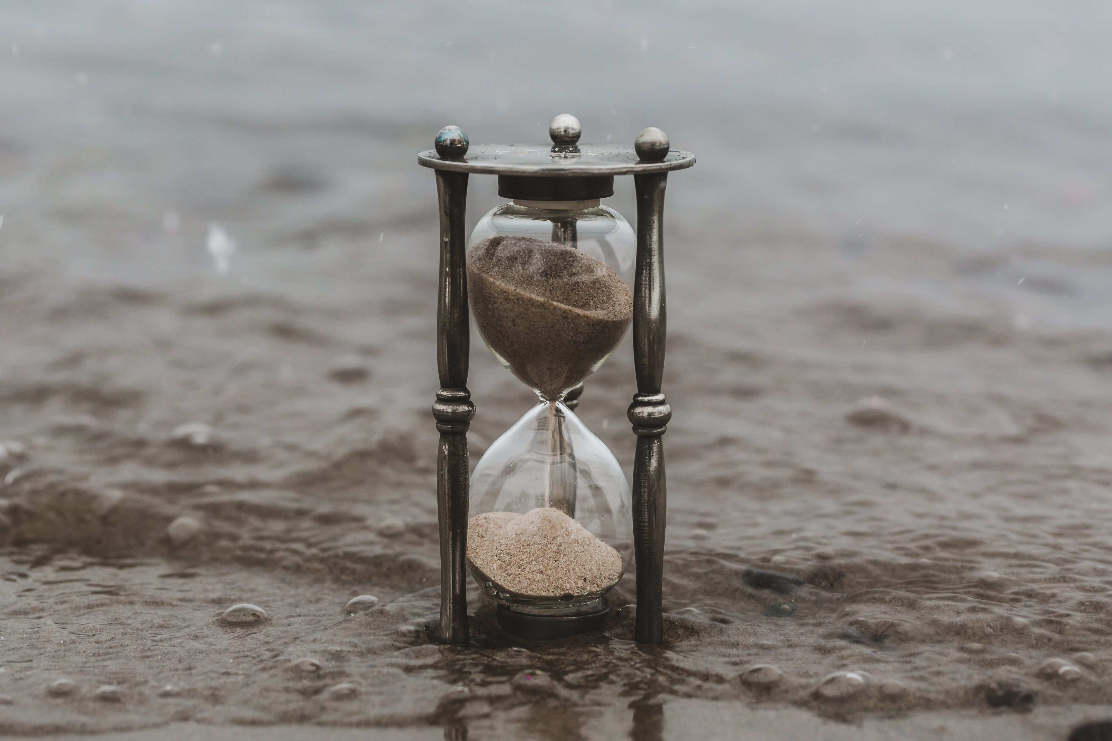 Eine antike Sanduhr mit metallischen Details steht auf einer nassen, sandigen Oberfläche. Der Sand in der oberen Hälfte der Sanduhr rieselt langsam in die untere Hälfte, während im Hintergrund verschwommen Wasser zu erkennen ist.