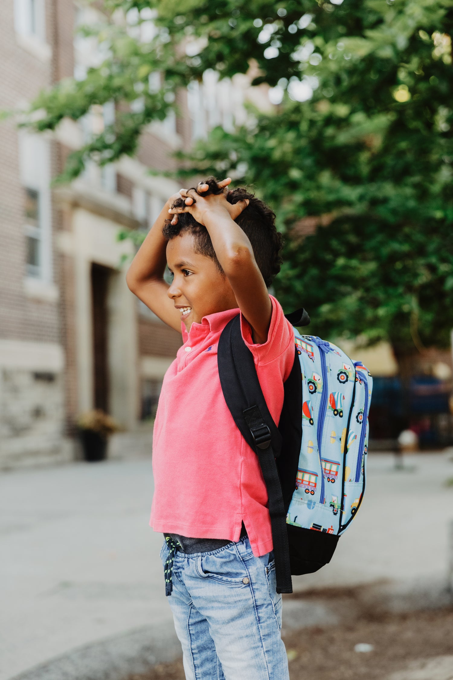 Ein Kind in einem leuchtend pinken T-Shirt steht auf einem Schulhof mit Bäumen im Hintergrund. Es trägt einen bunten Rucksack mit Motiven von Fahrzeugen und hat die Hände über den Kopf gelegt. Im Hintergrund sind unscharf ein Backsteingebäude und weitere Schulhofdetails zu erkennen.