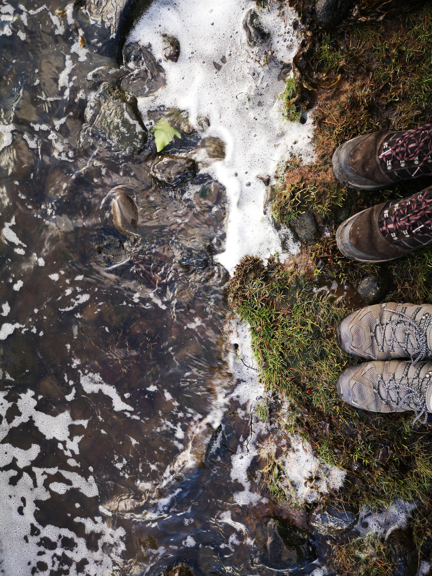Zwei Paar Schuhe stehen auf einer moosbewachsenen Uferkante neben einem flachen, mit Steinen bedeckten Gewässer. Schaum bildet sich an der Wasseroberfläche, und die Umgebung zeigt eine Mischung aus Gras, Erde und Steinen.