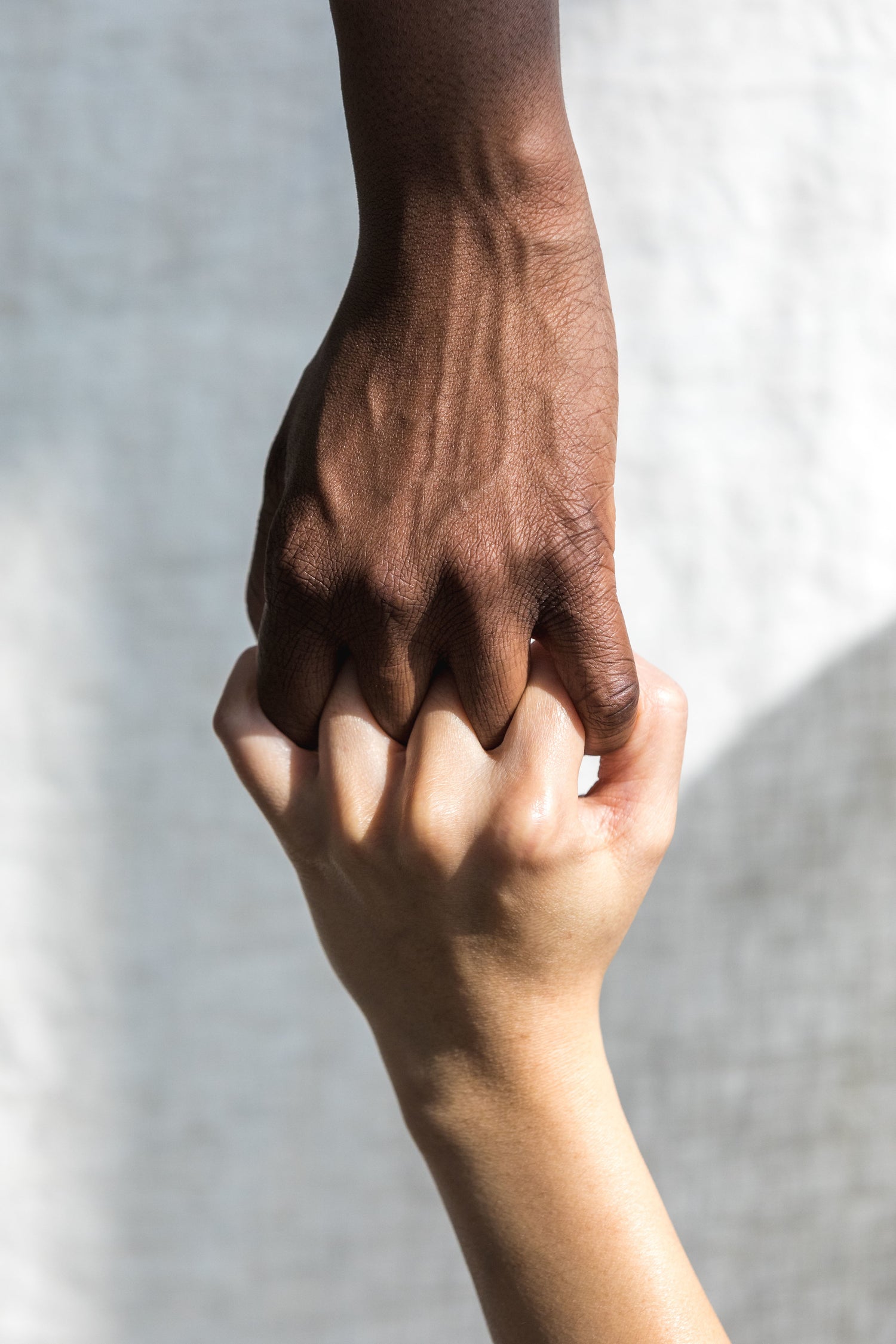 Zwei ineinander verschränkte Hände vor hellem Hintergrund: Eine dunklere Hand greift von oben eine hellere Hand, die von unten gehalten wird. Symbol für Zusammenhalt und Verbundenheit.