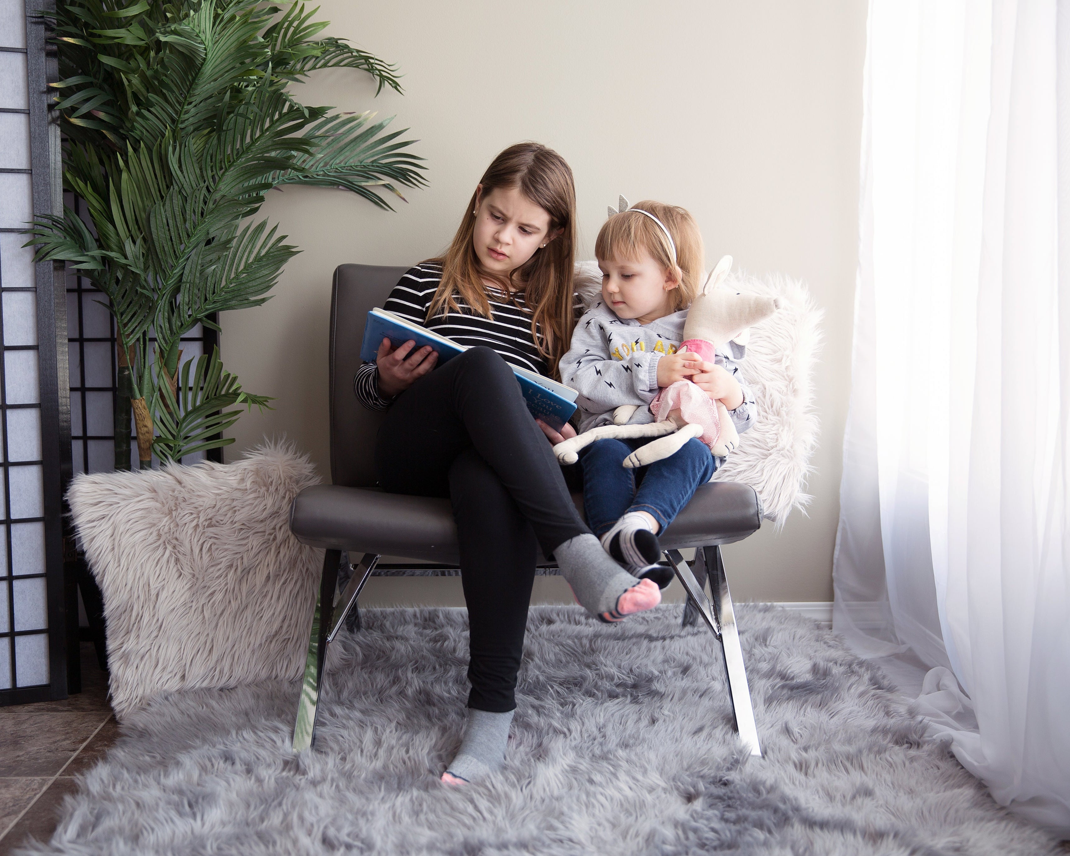 Zwei Kinder sitzen zusammen auf einem grauen Stuhl. Eines der Kinder hält ein Buch und liest, während das andere Kind ein Stofftier umarmt. Die Szene ist in einem gemütlichen Raum mit einem flauschigen Teppich, einer Zimmerpflanze und lichtdurchfluteten Vorhängen gestaltet.