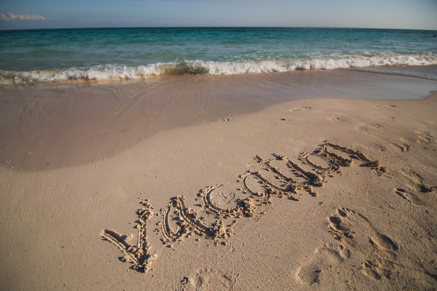 Strand mit sanften Wellen und klarem, türkisfarbenem Wasser im Hintergrund. Im Sand ist das Wort 'vacation' (Urlaub) handschriftlich geschrieben, umgeben von Fußspuren.