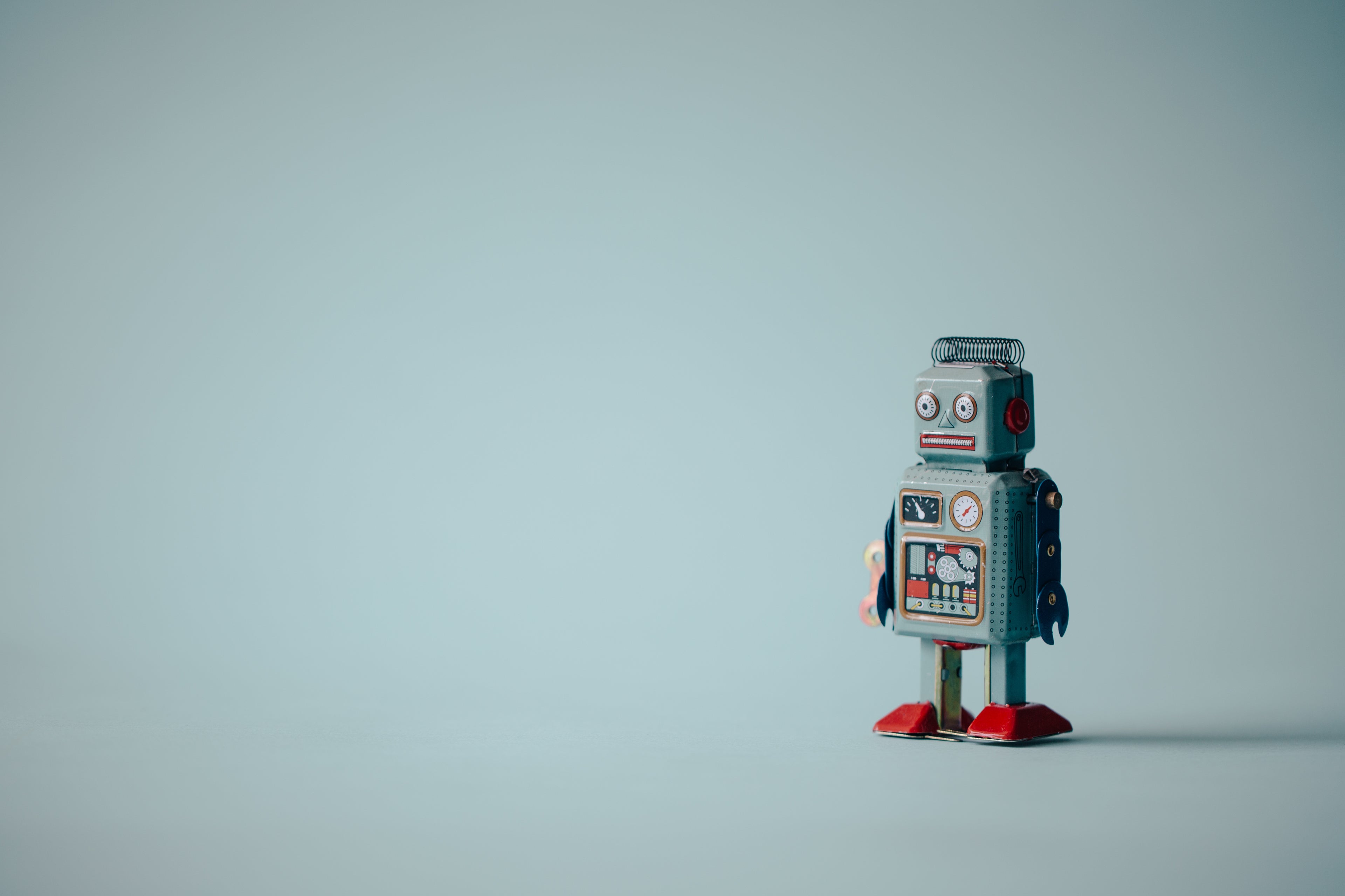 Spielzeug-Roboter aus Metall im Retro-Stil mit blauen Armen, roten Füßen und einem Zifferblatt auf der Brust. Der Roboter steht vor einem hellblauen Hintergrund.