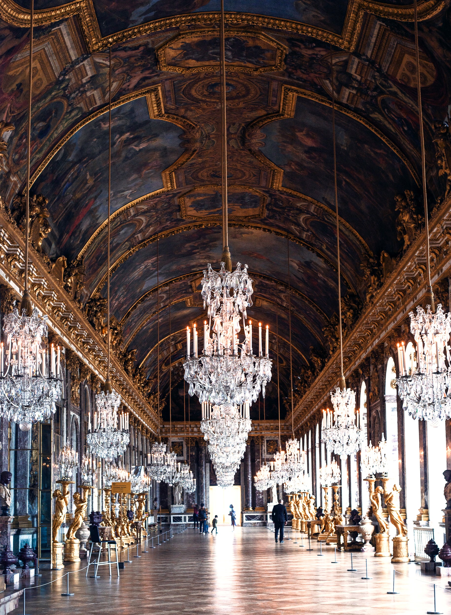 Prunkvoller Spiegelsaal mit einer hohen, kunstvoll bemalten Decke, zahlreichen Kristalllüstern und goldenen Statuen. Auf beiden Seiten des Saals befinden sich große Fenster, die viel Licht einlassen, und Spiegel, die den Raum optisch erweitern.