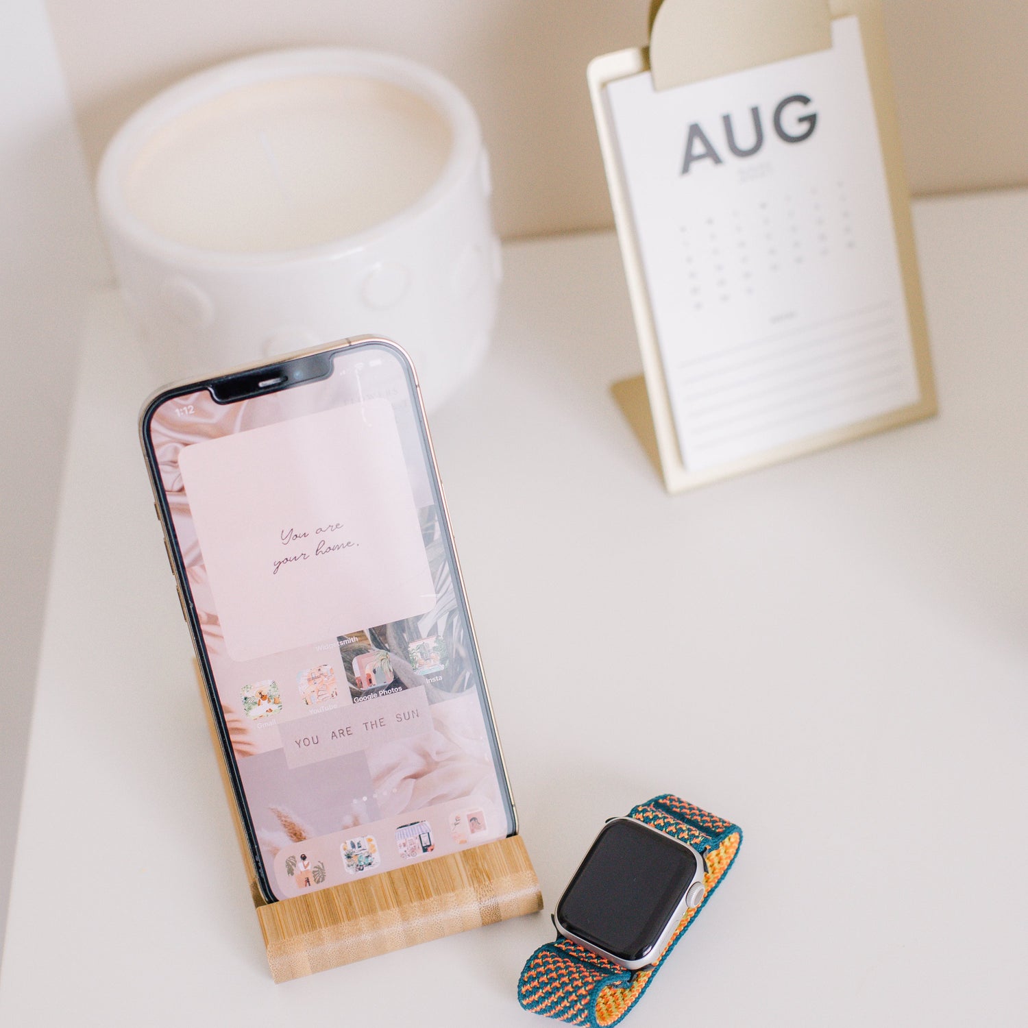Stillleben mit Smartphone im Holzständer, Smartwatch, Duftkerze und Tischkalender auf weißem Tisch.