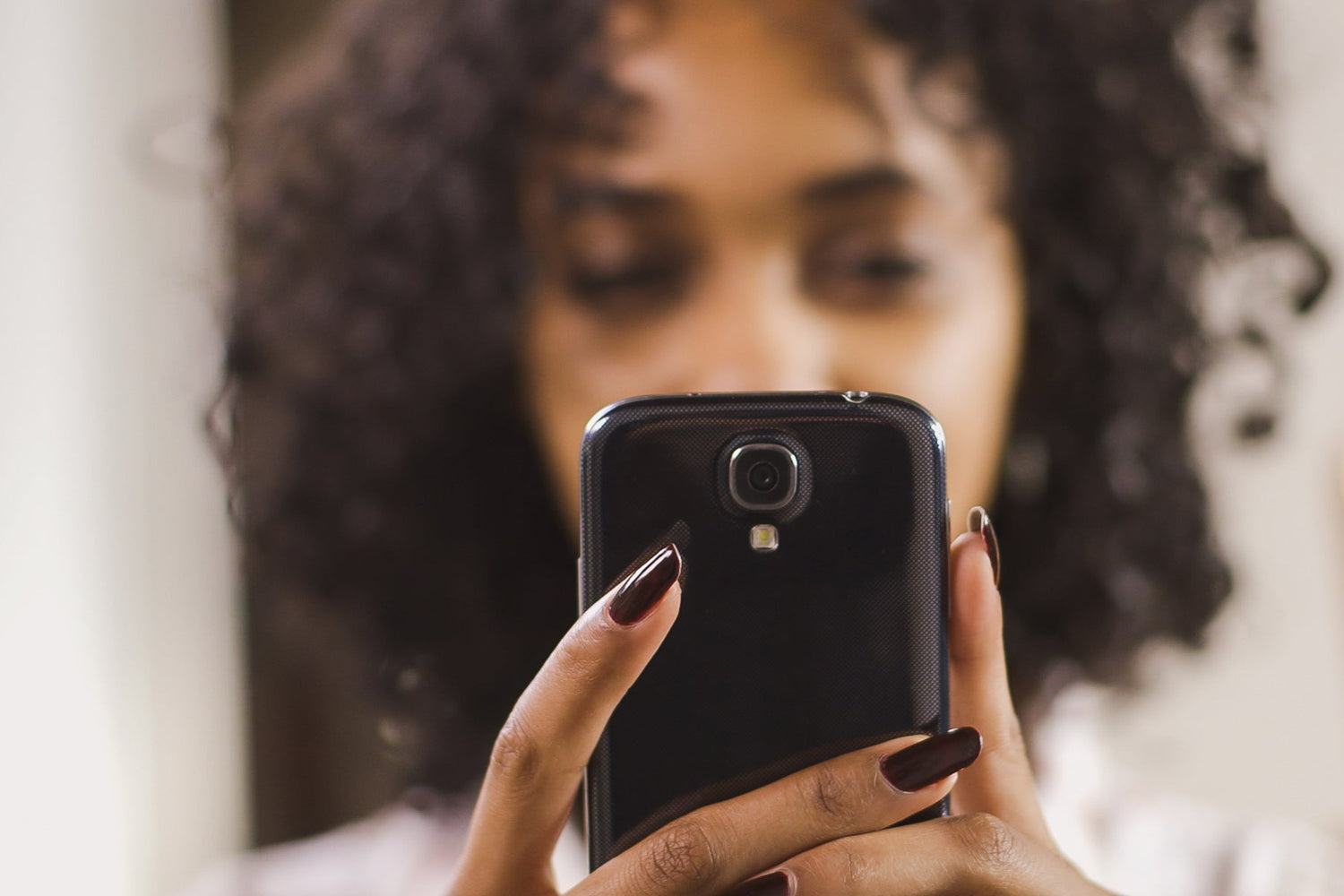 Person mit lockigem dunklem Haar hält ein Smartphone mit beiden Händen. Die Fingernägel sind dunkel lackiert. Im Hintergrund ist ein unscharfer, heller Raum zu sehen.