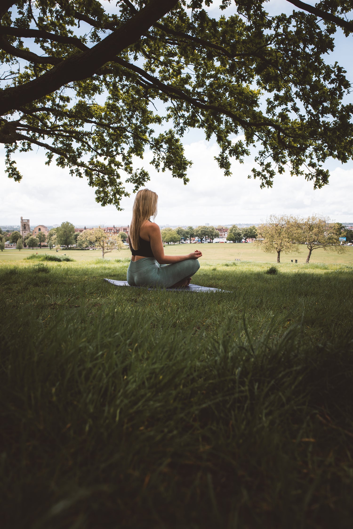 Eine Frau sitzt im Schneidersitz auf einer Yogamatte unter einem großen Baum in einer grünen Wiese und meditiert. Im Hintergrund ist eine weitläufige Landschaft mit Bäumen, Wiesen und einer kleinen Stadt mit Kirchturm zu sehen. Der Himmel ist leicht bewölkt, und die Atmosphäre wirkt ruhig und friedlich.
