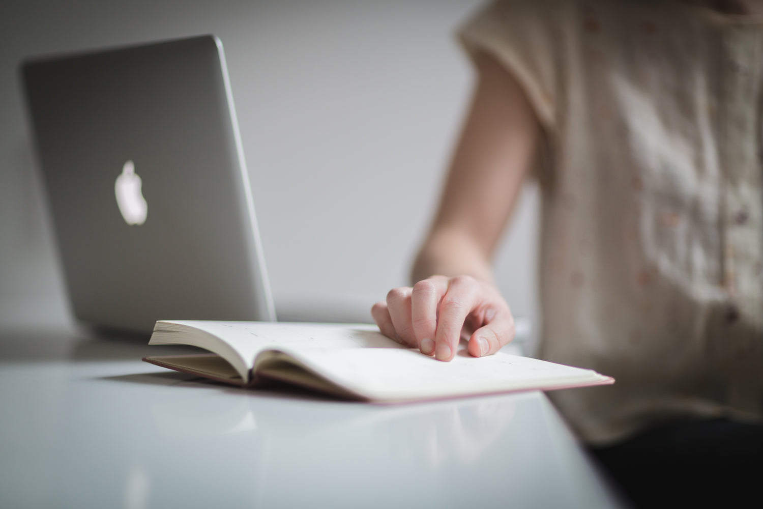 Eine Person sitzt an einem Schreibtisch, auf dem ein aufgeklapptes silbernes MacBook und ein aufgeschlagenes Notizbuch liegen. Die Person trägt ein helles Oberteil und zeigt mit dem Finger auf eine Stelle im Notizbuch, während der obere Teil ihres Körpers nicht vollständig im Bild ist.