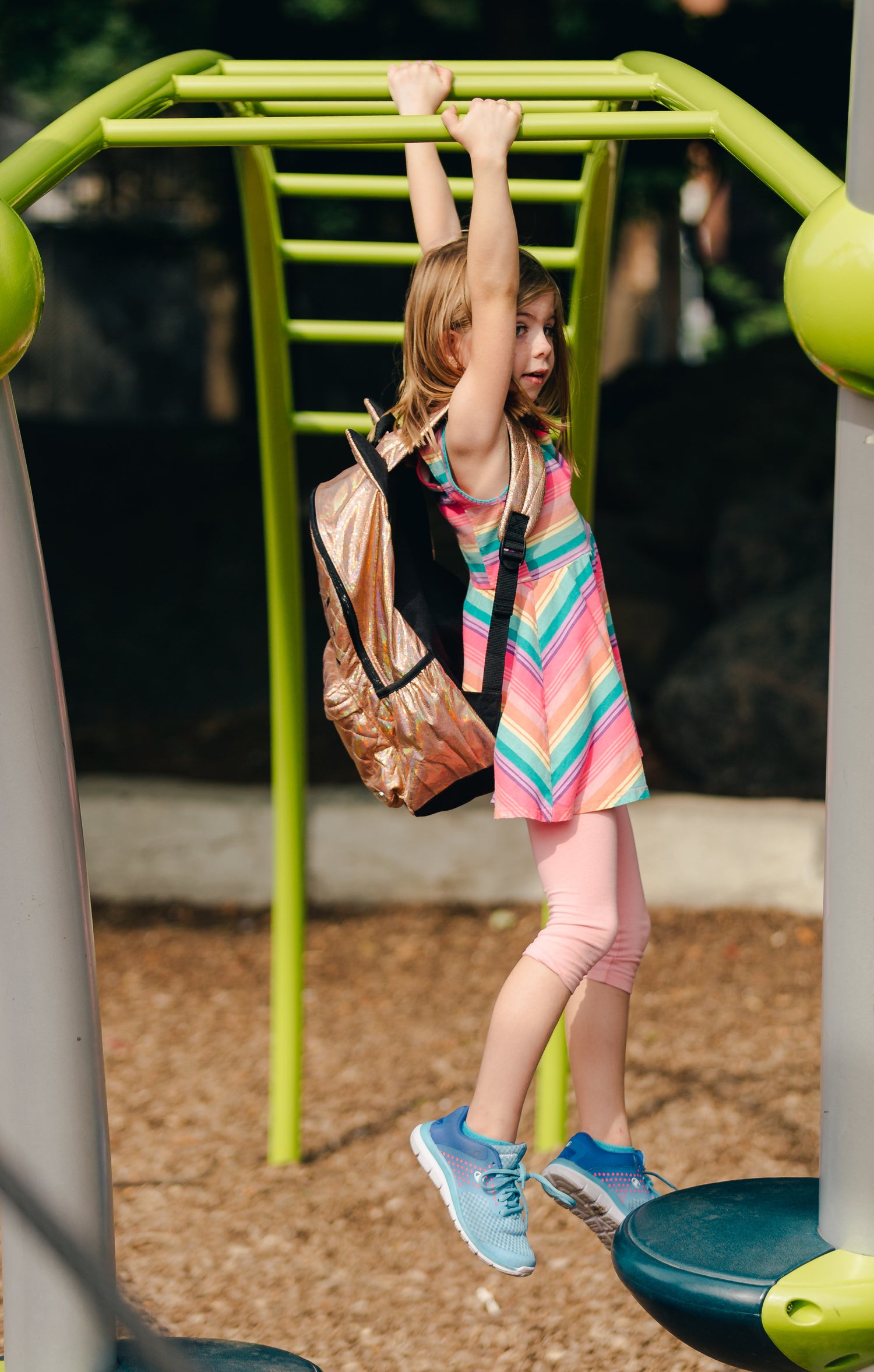 Ein Kind in bunter Kleidung schwingt sich an einer grünen Kletterleiter auf einem Spielplatz, während es einen goldenen Rucksack trägt. Der Boden ist mit Holzschnitzeln bedeckt, und im Hintergrund sind Bäume und schattige Bereiche zu sehen.
