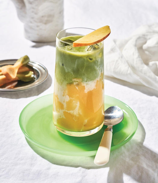 Ein Mango-Matcha-Latte im Glas mit Eiswürfeln, garniert mit einer Mango Scheibe. Die obere Hälfte des Getränkes ist Matcha-grün, die untere hälfte zeigt das gelb-orange einer Mango. 