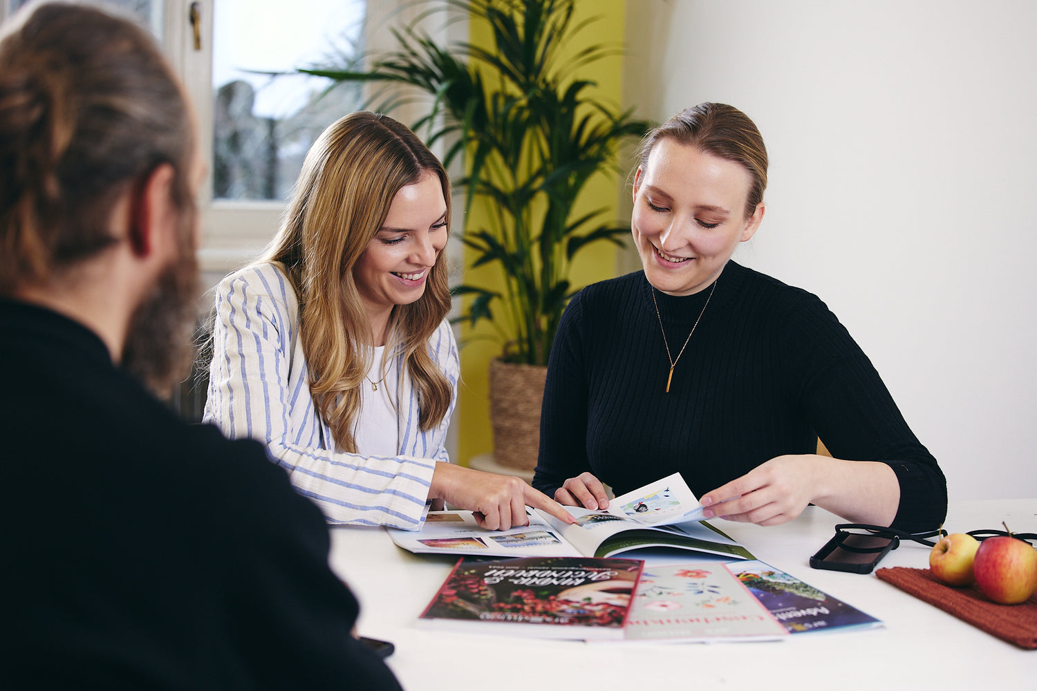 Zwei Personen sitzen an einem weißen Tisch und blättern gemeinsam in bunten Magazinen. Im Vordergrund ist eine weitere Person von hinten zu sehen. Auf dem Tisch liegen weitere Magazine, eine Brille und ein kleines Tablett mit Äpfeln. Im Hintergrund steht eine grüne Zimmerpflanze vor einer gelben Wand