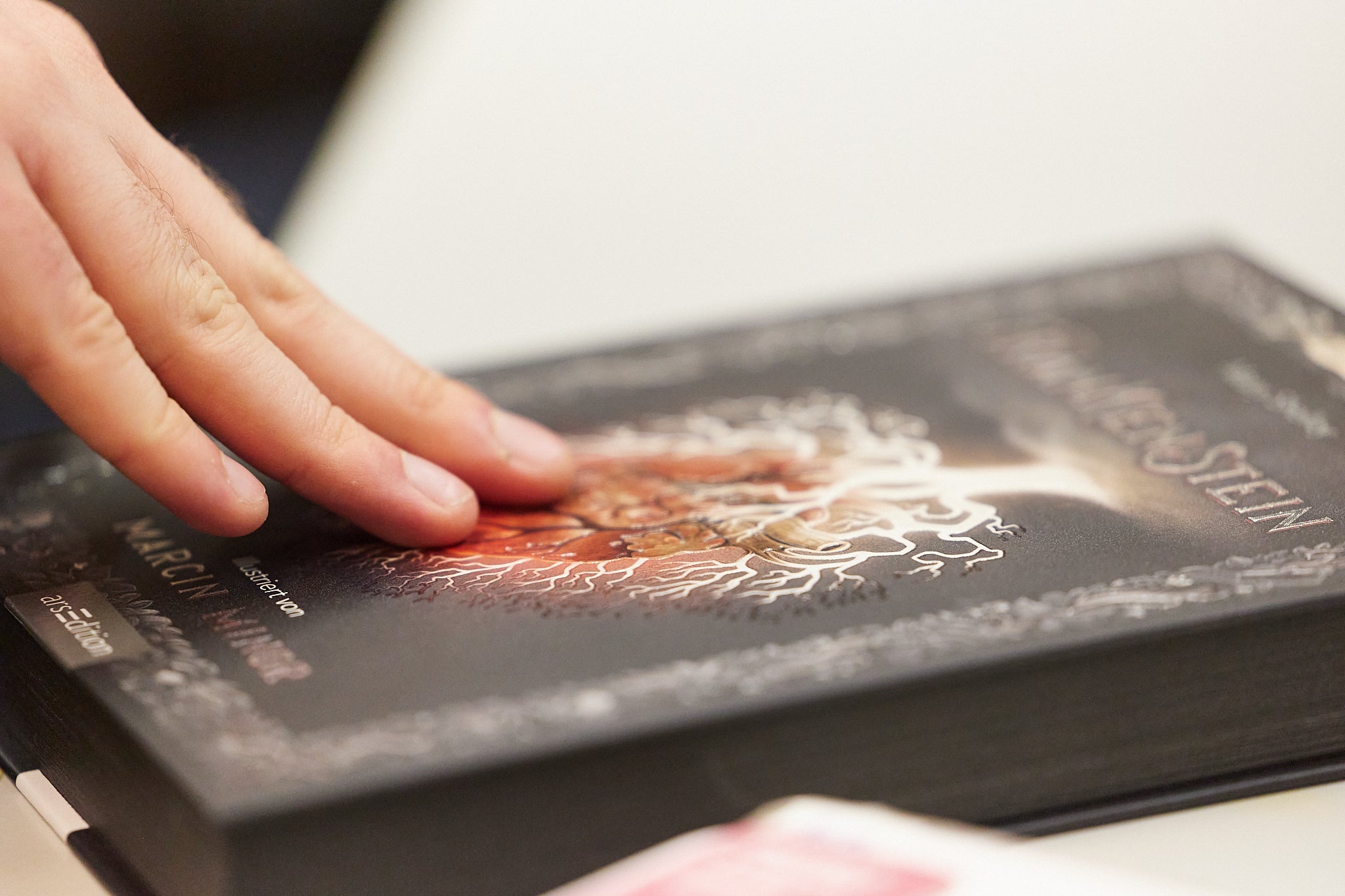 Eine Hand ruht auf dem Cover eines Buches mit dunklem Hintergrund und einem auffälligen, kunstvollen Motiv, das weiße und orangefarbene Details zeigt. Der Titel und der Name des Autors sind teilweise sichtbar.