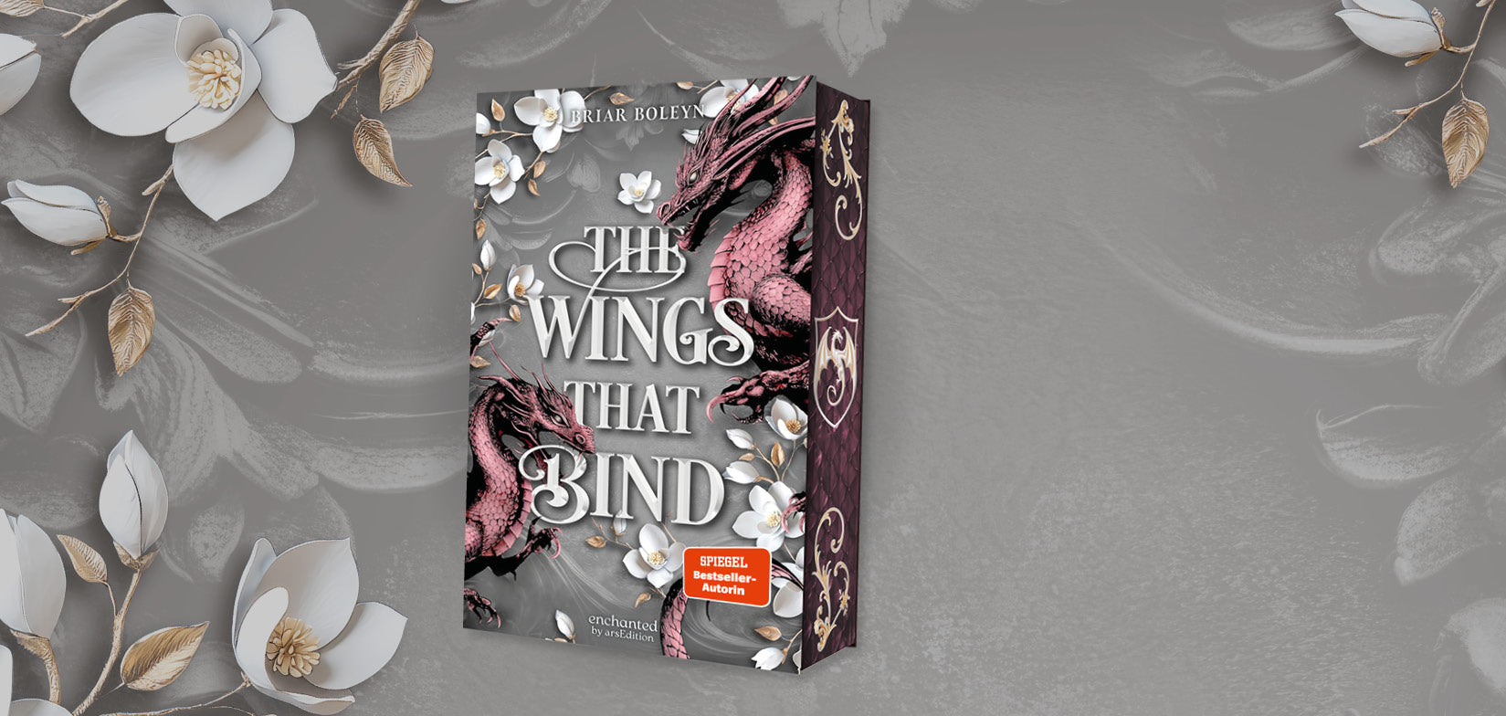 Breites Banner mit grauem, strukturiertem Hintergrund und weißen Blüten sowie Zweigen an den Rändern. In der Mitte steht ein aufgeschlagen wirkendes Buch mit dem Titel „The Wings That Bind“ von Briar Boleyn. Das Cover zeigt zwei rosafarbene Drachen, weiße Blumen und ein orangefarbenes Spiegel-Bestseller-Autorin-Siegel.