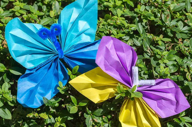 Zwei bunte Origami-Schmetterlinge aus Papier liegen auf grünem Blattwerk. Der linke Schmetterling ist blau mit einem Körper aus blauem Pfeifenreiniger, der rechte Schmetterling hat lila und gelbe Flügel mit einem weißen Pfeifenreiniger als Körper. Beide Schmetterlinge sind detailreich gefaltet und heben sich durch ihre leuchtenden Farben vom Hintergrund ab.