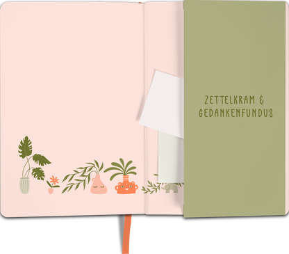 Gezeigt wird eine Innen- oder Zusatzansicht von „myNOTES Notizbuch A5: A home for good ideas“. Die linke Seite zeigt eine sanft rosa Hintergrundfarbe, verziert mit Illustrationen von Pflanzen in verschiedenen Töpfen. Auf der rechten Seite ist ein grüner Einleger mit dem Aufdruck „Zettelkrams &amp; Gedankenfundus“ in gut lesbarer, moderner Typografie. Die Farbgestaltung ist harmonisch und freundlich, ergänzt durch ein orangefarbenes Band, das als Lesezeichen dient.