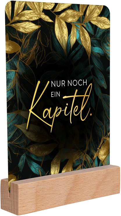 Gezeigt wird eine Innen- oder Zusatzansicht von „myNOTES Papeterie: Kartenbox Romance“. Die Ansicht präsentiert eine elegante Tafel, die mit goldenen und blauen Blättern auf einem tiefschwarzen Hintergrund geschmückt ist. In der Mitte heben sich die Worte „Nur noch ein Kapitel.“ in geschwungener, goldener Schrift hervor. Die Tafel ruht auf einem einfachen Holzständer, der für eine harmonische Gesamtkomposition sorgt und stilvolle Akzente setzt.