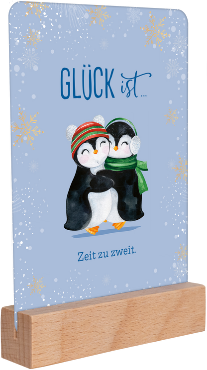 Gezeigt wird eine Innen- oder Zusatzansicht von „Weihnachtspost für dich und mich“. Auf einem hellblauen Hintergrund sind zwei liebevoll illustrierte Pinguine zu sehen, die sich umarmen. Der Text „GLÜCK ist…“ in verspielter Schrift leuchtet in Blau, während der Satz „Zeit zu zweit.“ in einer klaren, gut lesbaren Schrift darunter steht. Der untere Bereich wird von einem naturfarbenen Holzsockel ergänzt, der das Bild harmonisch abrundet. Goldene Schneeflocken verleihen der Szene...