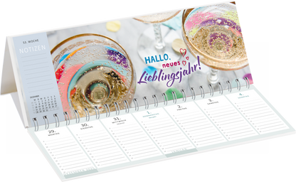 Gezeigt wird eine Innen- oder Zusatzansicht von „Tischkalender Heute ist mein Lieblingstag! 2026“. Die Seite präsentiert einen festlichen Hintergrund mit Gläsern, die mit prickelndem Getränk gefüllt sind, sowie bunten Konfetti-Elementen. Der ansprechende Schriftzug „HALLO, neues Lieblingsjahr!“ in lebhaften Farben steht im Vordergrund. Darunter sind die Kalenderwochen mit ausreichend Platz für Notizen, die in klaren, strukturierten Linien dargestellt sind. Die Farbpalette ist hell...