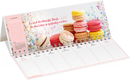 Gezeigt wird eine Innen- oder Zusatzansicht von „Tischkalender Glücksmomente 2026“. Der Kalender präsentiert eine ansprechende Illustration von bunten Makarons auf einem blütenhaften Hintergrund. Oben links befindet sich ein Bereich für Notizen, im sanften Rosaton, mit klaren, gut lesbaren Zahlen und Monatsangaben. Die zentrale Aussage „Es sind die kleinsten Dinge, die das Leben so großartig machen“ ist in einer verspielten, handgeschriebenen Schriftart in einem warmen Rotton...