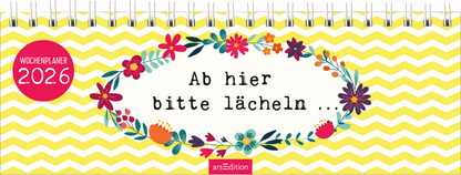 Das Cover zeigt einen bunten Wochenplaner mit einem fröhlichen, gelben Wellenmuster im Hintergrund. In der Mitte umrahmt eine blumige Gestaltung den Text „Ab hier bitte lächeln …“, der in kräftiger, schwarzer Schrift geschrieben ist. Oben links befindet sich ein roter Button mit dem Text „WOCHENPLANER 2026“. Ganz unten mittig steht der Logoschriftzug von arsEdition.