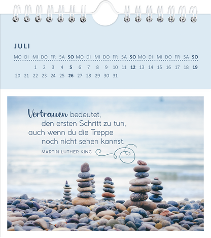 Gezeigt wird eine Innen- oder Zusatzansicht von „Wandkalender Gelassenheit für dich 2026“. Die Ansicht zeigt den Monat Juli, gekennzeichnet durch eine helle, sanfte Farbpalette in Blau- und Grautönen. Die oberste Zeile enthält den Monatsnamen, während der Kalender in einem klaren und leserlichen Typografie-Design die Wochentage anzeigt. Im Vordergrund steht ein inspirierendes Zitat von Martin Luther King, umgeben von einer Fotografie von sorgfältig gestapelten Steinen am Wasser, was...