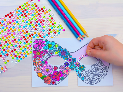 Gezeigt wird eine Innen- oder Zusatzansicht von „Diamantmasken – Glitzerspaß“. Die Abbildung zeigt eine bunte, bereits teilweise kolorierte Maske mit floralen Motiven in kräftigen Farben wie Pink, Blau und Gelb. Im Hintergrund sind farbige Buntstifte und ein Bogen mit glitzernden Stickern zu sehen, die eine Vielzahl von Farben bieten. Eine Hand platziert gerade einen glitzernden Sticker auf die Maske, was die Kreativität und den Spaß am Basteln unterstreicht.