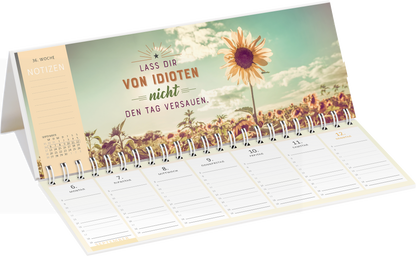 Gezeigt wird eine Innen- oder Zusatzansicht von „Tischkalender Meine Motivation &amp; ich leben gerade getrennt. 2027“. Die Ansicht zeigt eine Spiralbindung und einen hellen Hintergrund mit einem großen Sonnenblumenmotiv. Darauf steht in kräftigen, roten Buchstaben „LASS DIR VON IDIOTEN nicht DEN TAGVERSAUEN“. Unterhalb befinden sich Wochenkalendertage mit ausreichend Platz für Notizen, umgeben von einem harmonischen Farbschema in sanften Pastelltönen. Die Typografie ist modern und...
