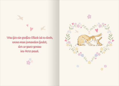 Gezeigt wird eine Innen- oder Zusatzansicht von „Buchkalender Weißt du eigentlich, wie lieb ich dich hab? 2027“. Auf einer sanften, cremefarbenen Hintergrund sind zwei niedliche Hasen in einem Herz aus floralen Elementen abgebildet. Umrahmt von zarten Blumen und kleinen Herzen strahlt die Illustration Wärme aus. Der Text in kräftigem Rot hebt sich ab und lautet: „Was für ein großes Glück ist es doch, wenn man jemanden findet, der so ganz genau ins Herz passt.“ Die Schriftart ist...