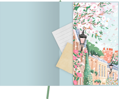 Gezeigt wird eine Innen- oder Zusatzansicht von „myNOTES Papeterie: Notizbuch A4 Simply, Katy – Spring Lane“. Die Innenseite ist in sanften Blau- und Pastelltönen gehalten. Auf der rechten Seite verziert eine florale Illustration mit rosa Blüten und Straßenlaternen eine charmante Gebäudefront. Links befinden sich einfache, liniierte Seiten und ein beige Umschlag. Die Verwendung von verspielter Typografie ergänzt das harmonische Design und schafft ein einladendes Ambiente.