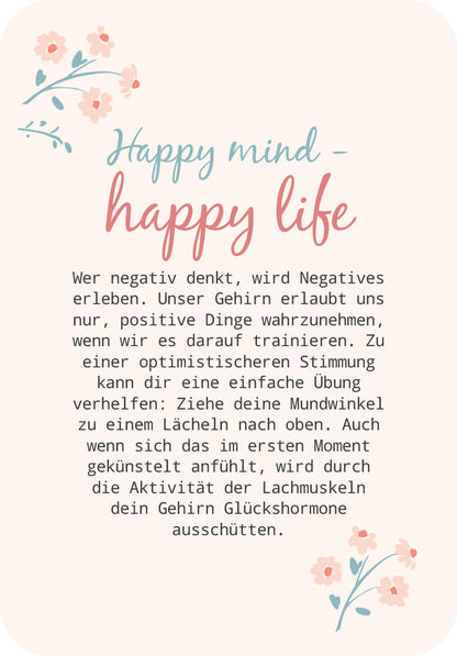 Gezeigt wird eine Innen- oder Zusatzansicht von „Glücksmantras für dich“. Die Seite präsentiert den Titel „Happy mind - happy life“ in einer verspielten, zweigeteilten Typografie: der erste Teil in sanftem Blau, der zweite in einem warmen Rosa. Darunter folgt ein erläuternder Text in einer klaren, gut lesbaren Schriftgröße auf hellem Hintergrund, umrahmt von zarten Blumenillustrationen in Pastelltönen, die eine positive und einladende Stimmung erzeugen.