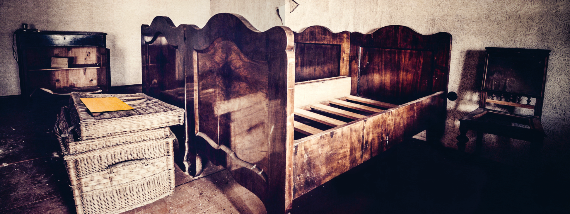 Gezeigt wird eine Innenansicht von „Escape Room. Das Hotel der tausend Augen“. Der Raum präsentiert sich mit einem rustikalen Holzbett mit geschwungenen Kopf- und Fußteilen in dunklen Brauntönen. Links befinden sich zwei Korbbehälter, während ein kleiner Holzschrank im Hintergrund steht. Auf dem Bett liegt ein gelbes Blatt. Die Wände sind in neutralen Farben gehalten, was eine atmosphärische, geheimnisvolle Stimmung erzeugt.