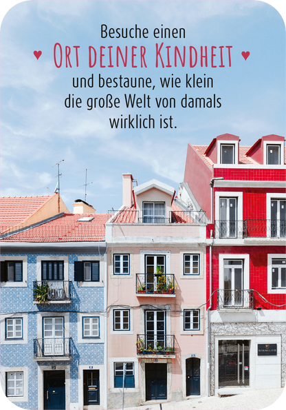 Gezeigt wird eine Innen- oder Zusatzansicht von „99 herrliche Auszeiten für zwischendurch“. Die Ansicht zeigt bunte, sanierte Häuserfassaden in Pastellfarben, darunter Blau, Rot und Weiß. Über den Gebäuden ist der handschriftliche Text „Besuche einen Ort deiner Kindheit und bestaune, wie klein die große Welt von damals wirklich ist“ in dunkler Schrift auf hellem Hintergrund platziert. Die Schrift ist verspielt und gut lesbar, was eine freundliche Atmosphäre vermittelt.