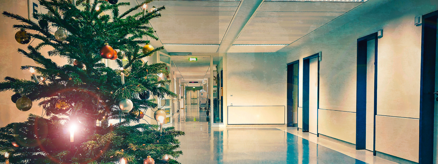 Gezeigt wird eine Innenansicht von „Escape Room. Patient 13“. In der Szene ist ein heller Krankenhausflur zu sehen, links steht ein geschmückter Weihnachtsbaum mit bunten Kugeln in Gold, Silber und verschiedenen Farben. Der Boden ist in einem sanften Blau gehalten, die Wände sind hell und schlicht. An der rechten Seite befinden sich mehrere Türen, die durch dunkle Rahmen abgehoben sind. Die Beleuchtung des Flurs ist neutral und sorgt für eine ruhige Atmosphäre.