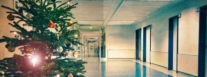 Gezeigt wird eine Innenansicht von „Escape Room. Patient 13“. In der Szene ist ein heller Krankenhausflur zu sehen, links steht ein geschmückter Weihnachtsbaum mit bunten Kugeln in Gold, Silber und verschiedenen Farben. Der Boden ist in einem sanften Blau gehalten, die Wände sind hell und schlicht. An der rechten Seite befinden sich mehrere Türen, die durch dunkle Rahmen abgehoben sind. Die Beleuchtung des Flurs ist neutral und sorgt für eine ruhige Atmosphäre.