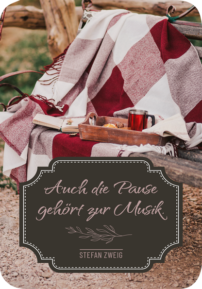 Gezeigt wird eine Innen- oder Zusatzansicht von „Weisheiten für die Seele“. Auf einem rustikalen Holzstuhl liegt eine gemusterte Decke in Rot- und Grautönen. Neben der Decke steht eine kleine Holzschale mit Snacks und einem Getränk in einem Glas. Im Vordergrund befindet sich ein elegantes, geschwungenes Textfeld in dunklem Braun, auf dem in einer verspielten Schrift „Auch die Pause gehört zur Musik“ und darunter „Stefan Zweig“ zu lesen ist.