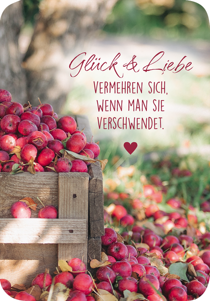 Gezeigt wird eine Innen- oder Zusatzansicht von „Weisheiten für die Seele“. Im Vordergrund befindet sich eine Holzkiste, gefüllt mit leuchtend roten Äpfeln. Der Hintergrund zeigt unscharf grünes Laub und einen baumartigen Schatten. In elegant geschwungener, roter Schrift steht der Text „Glück &amp; Liebe“ über einem Zitat, das in einer klaren, schwarzen Schrift gewählt ist. Ein rotes Herz ergänzt das Design. Die Gesamtkomposition wirkt warm und einladend.