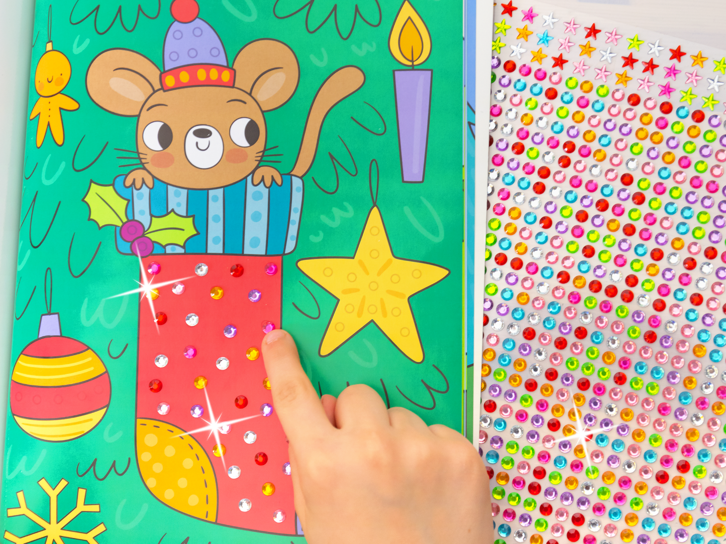 Es wird eine Innenseite des Buches „Mein Diamant-Stickerheft – Wintertraum“ in Aktion gezeigt: Eine Kinderhand ragt von unten in das horizontale Bild rein und klebt soeben einen Diamantsticker auf die Innenseite. Auf dem Bild sind bereits alle Felder mit Diamanten geschmückt und glänzt daher wunderschön. Unterhalb des Heftes ragt der Stickerbogen hervor und zeigt eine große Vielzahl an bunten Diamantstickern.